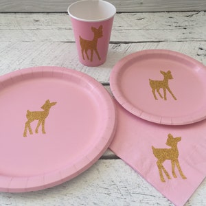 May include: Pink paper plates and cups with gold glitter deer silhouettes. The plates and cups are part of a party supply set.