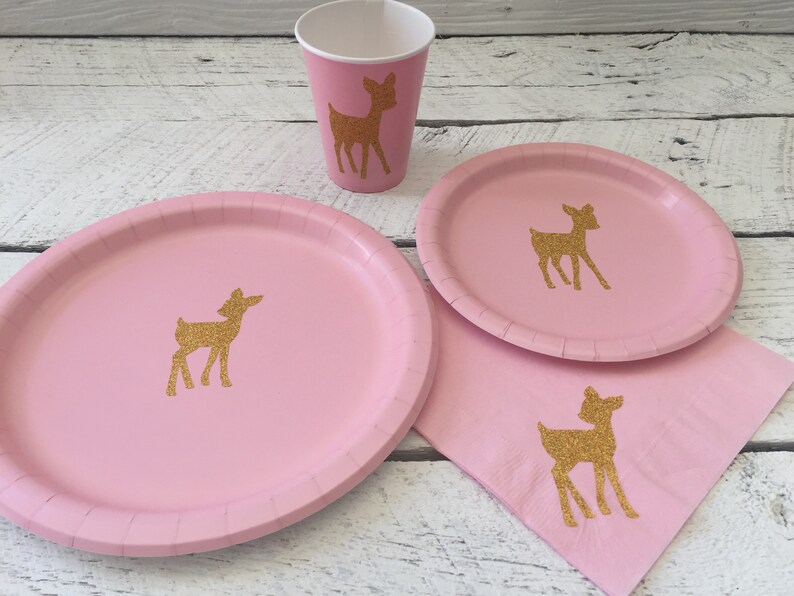 May include: Pink paper plates and cups with gold glitter deer silhouettes. The plates and cups are part of a party supply set.