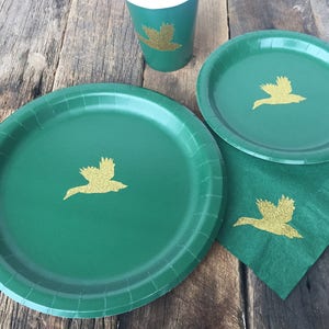 May include: Three green paper plates with gold glitter duck silhouettes. The plates are on a wooden surface.