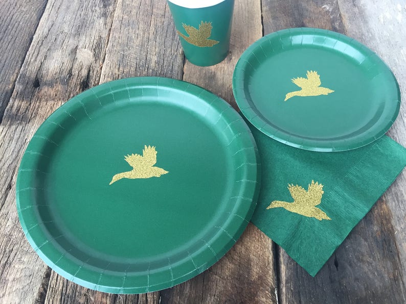 May include: Three green paper plates with gold glitter duck silhouettes. The plates are on a wooden surface.
