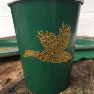 May include: A green paper cup with a gold glitter flying duck design. The cup has a white rim.