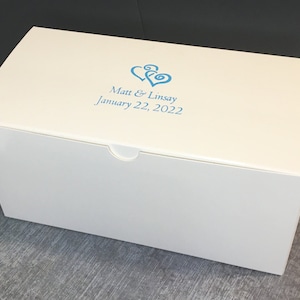 May include: A rectangular white box with a hinged lid featuring a blue graphic of two interlocking hearts and the printed text Matt & Linsay January 22, 2022. The box has a smooth finish and a small semi-circular cutout on the front edge for opening.