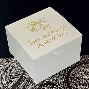 May include: White favor box with a gold design of wedding bells and the text "Steven and Carolyn April 7th, 1973".