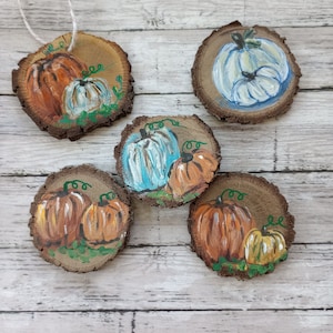 May include: Five wooden round ornaments with painted pumpkins. The ornaments are painted with orange, white, and blue pumpkins. Some of the pumpkins have green stems and leaves.