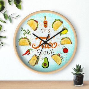 May include: A round wooden wall clock with a blue background and a white design. The clock features a variety of illustrations of tacos, avocados, chili peppers, and a bottle of hot sauce. The clock face reads "It's Taco Clock".