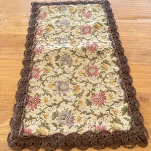 May include: A rectangular table runner with a floral pattern. The runner has a cream-coloured background with pink, yellow, and purple floral designs. It is edged with a dark brown, scalloped lace trim, adding a decorative touch to the table decor.