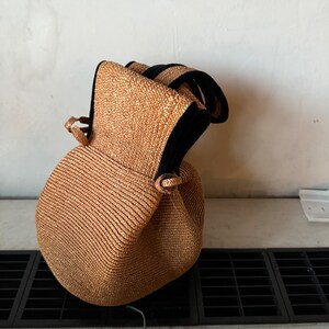 May include: A brown woven straw bag with black trim and a drawstring closure. The bag is sitting on a white surface with a black grate.