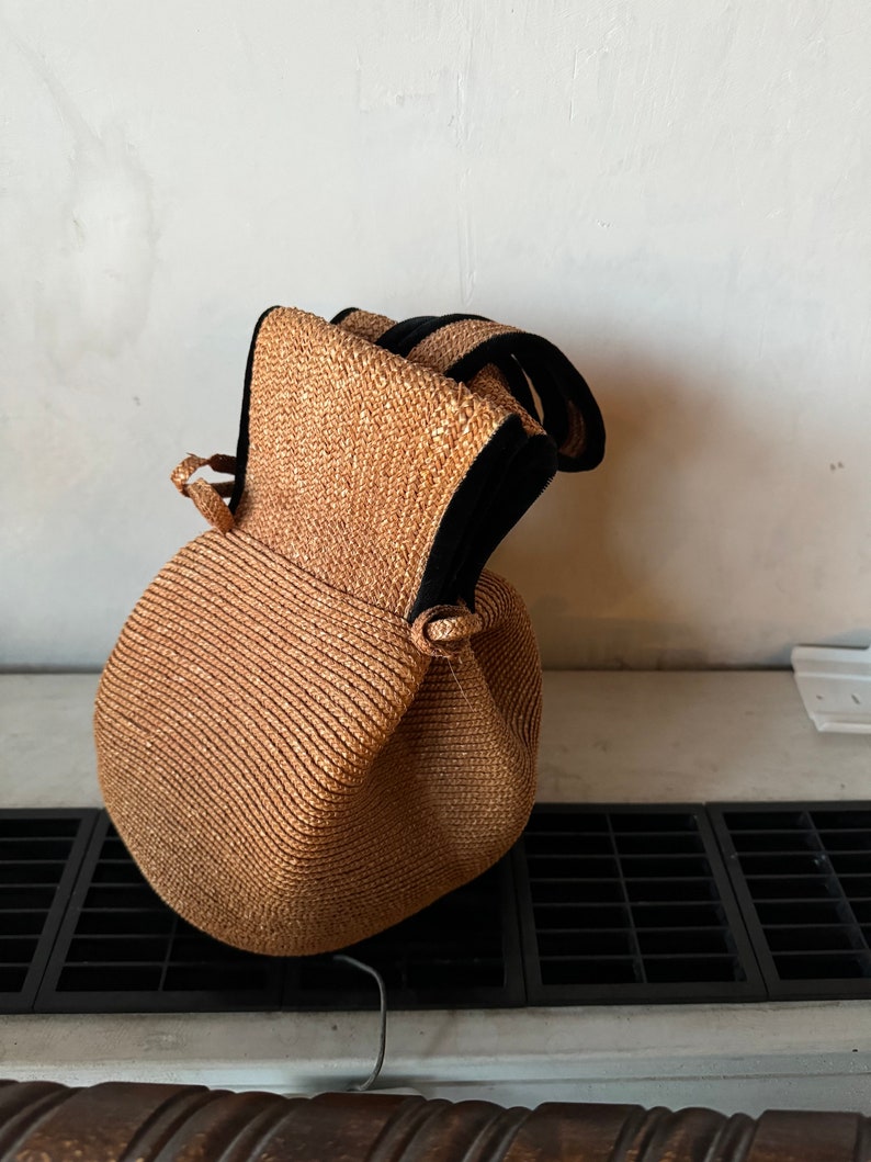 May include: A brown woven straw bag with black trim and a drawstring closure. The bag is sitting on a white surface with a black grate.