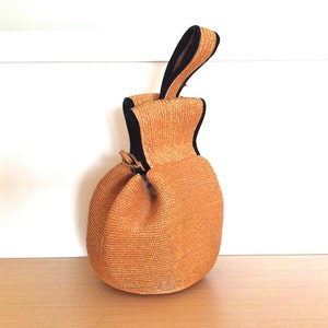 May include: A small, round, orange straw bag with a black handle. The bag has a woven texture and a drawstring closure.