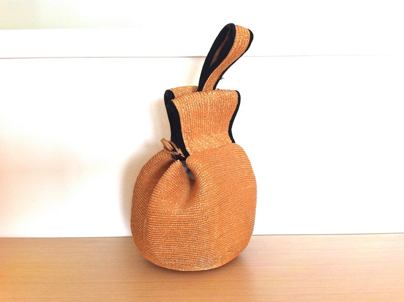 May include: A small, round, orange straw bag with a black handle. The bag has a woven texture and a drawstring closure.