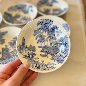 May include: A set of four white ceramic bowls with blue and white floral designs. The bowls feature a landscape scene with trees, a house, and a boat.