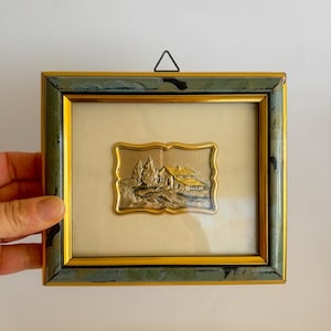 May include: A framed metal relief showing a rural landscape with trees and a house. The artwork is set within a gold-lined frame with a green and gold outer frame. The piece is designed to be hung on a wall.