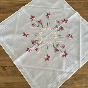 May include: White square tablecloth with a delicate floral embroidery design. The embroidery features pink and white flowers in a circular pattern.