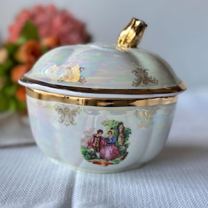 Peut inclure: Un récipient en céramique blanche en forme de citrouille avec un couvercle et des garnitures dorés. Le récipient présente une scène peinte d'un couple dans un jardin.