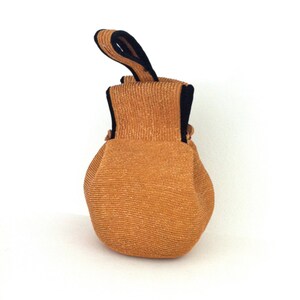 May include: A small, round, brown woven bag with a black fabric handle. The bag is made of a textured material and has a slightly rounded shape.