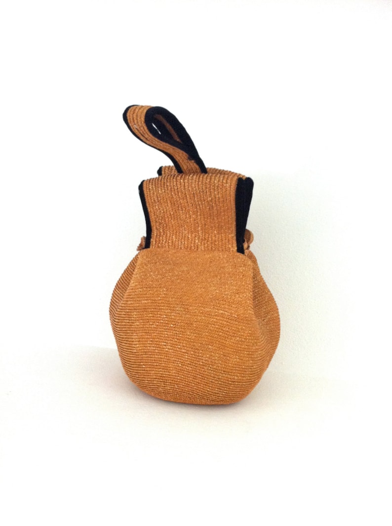 May include: A small, round, brown woven bag with a black fabric handle. The bag is made of a textured material and has a slightly rounded shape.