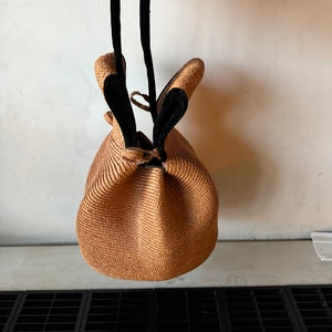 May include: A brown woven straw bag with black leather straps. The bag is round and has a drawstring closure.