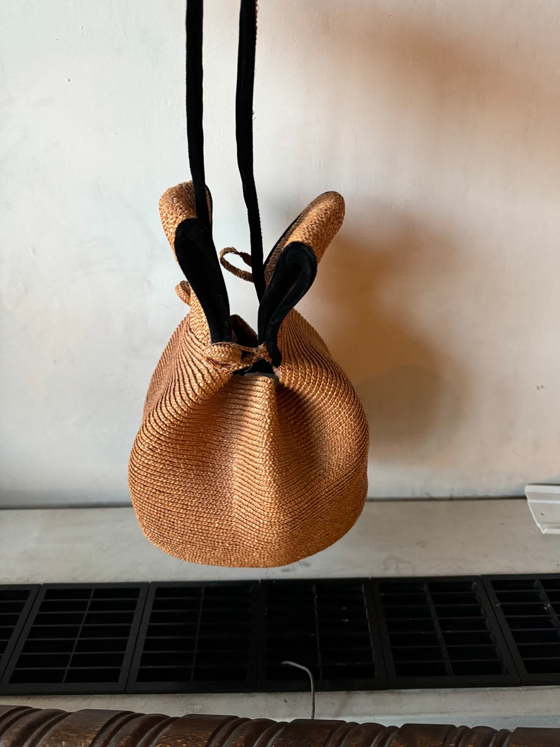 May include: A brown woven straw bag with black leather straps. The bag is round and has a drawstring closure.