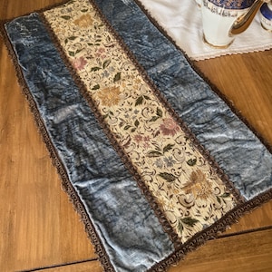 May include: A rectangular table runner with a central floral patterned panel flanked by blue velvet sections. The runner is trimmed with a brown, decorative border. The floral pattern includes gold, pink, and green elements.