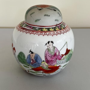 Peut inclure: Un pot en porcelaine blanche avec un couvercle, décoré d'une scène colorée de personnes pêchant. Le pot a un bord rouge et un fond blanc.