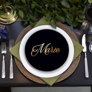 K&ouml;nnte beinhalten: Eine formelle Tischdekoration mit einem schwarzen Teller mit dem Namen "Marie" in goldener Schrift. Der Teller steht auf einem wei&szlig;en Teller, einer gr&uuml;nen Serviette und einem gewebten Platzset. Besteck und ein Weinglas sind ebenfalls vorhanden.