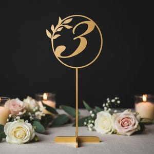 Puede incluir: Un número de mesa dorado, el número 3, con un diseño circular y una ramita de hojas. El número está sobre un soporte con base en forma de cruz. El fondo incluye velas y flores, ideal para una boda o evento.