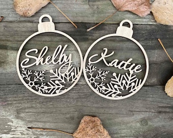 May include: Two wooden Christmas ornaments with snowflake designs. The ornaments are personalized with the names "Shelly" and "Katie".
