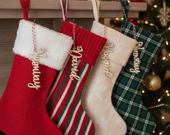 May include: Four Christmas stockings hang from a mantel. The stockings are red, green and white striped, cream with snowflakes, and green plaid. Each stocking has a wooden name tag and a red and white hanging loop.