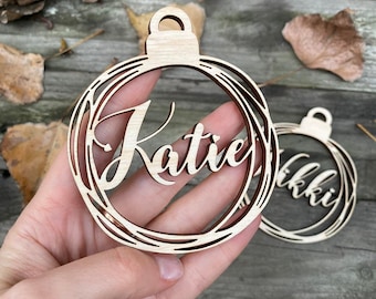 May include: Two wooden Christmas ornaments with the names "Katie" and "Nikki" cut out in a decorative font. The ornaments are a light brown color and have a circular shape.