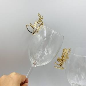 May include: Two clear wine glasses with gold-coloured name tags that read "Cheryl" and "Valerie". The tags are attached to the rims of the glasses. The background is plain white.