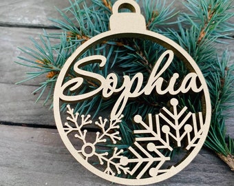 May include: A wooden Christmas ornament shaped like a ball with the name "Sophia" cut out in the center. The ornament is decorated with snowflakes and pine branches.