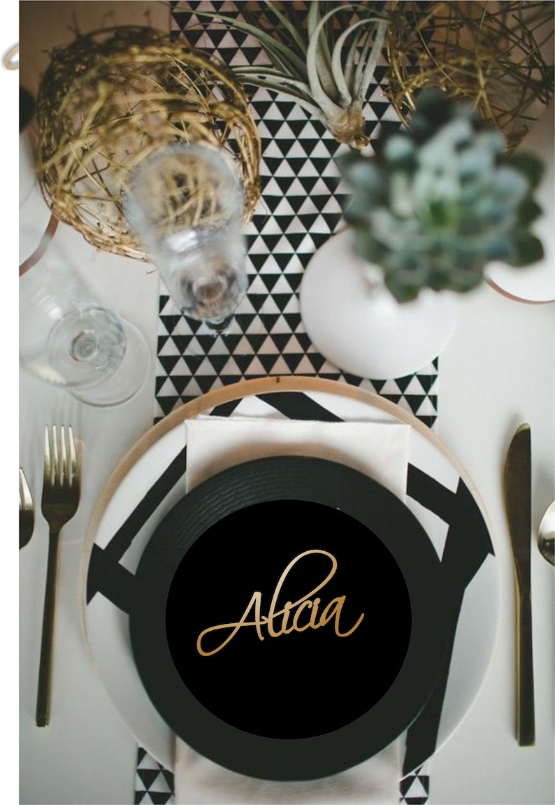 K&ouml;nnte beinhalten: Eine Tischdekoration mit einem schwarzen Teller mit dem Namen "Alicia" in goldener Schrift. Das Arrangement umfasst goldenes Besteck, einen wei&szlig;en Teller mit einem schwarz-wei&szlig;en geometrischen Design und dekorative Elemente.