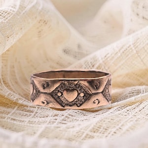 May include: A copper-toned metal ring featuring a repeating pattern of embossed diamonds, each containing a small heart shape at the center, surrounded by textured detailing and small circular accents along the band's outer surface.