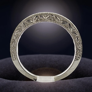 May include: A silver ring with an intricate floral pattern around the band. The ring is set against a dark blue background, with a soft light illuminating the ring's details. The ring's design features small, repeating leaf-like shapes.