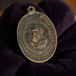 May include: Oval-shaped antique bronze pendant featuring a portrait of a bearded man. The pendant has a loop at the top for hanging and is surrounded by a border of stars and decorative elements. The portrait is in a central oval.