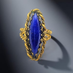 May include: A gold-toned ring featuring a large, elongated, deep blue gemstone. The gemstone is set within a detailed, twisted rope-like frame, with additional small decorative elements around the stone. The ring has an antique or vintage aesthetic.