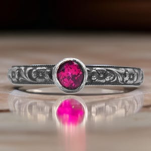 May include: A silver ring with a pink gemstone set in a bezel setting. The band is decorated with an engraved floral design.