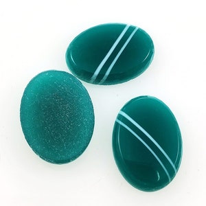 May include: Three oval-shaped teal green glass tiles with white stripes. Two of the tiles have a smooth, glossy finish, while the third tile has a textured, frosted finish.