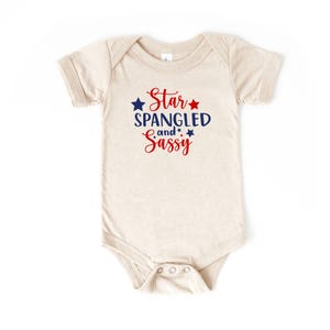 Może przedstawiać: Beżowe body niemowlęce z napisem "Star Spangled and Sassy" w kolorze czerwonym i niebieskim. Wzór zawiera gwiazdki. Body ma krótkie rękawy i zapięcie na zatrzaski.