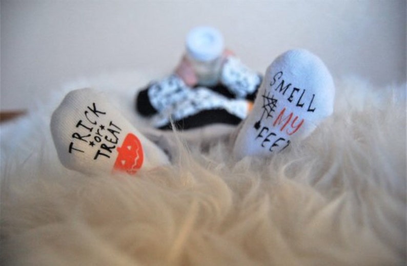 May include: A pair of white socks with Halloween-themed text. One sock says "Trick or Treat" with a small orange pumpkin graphic. The other sock says "Smell My Feet?" with a small spider web graphic.