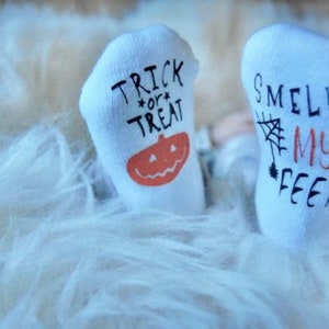 May include: A pair of white baby socks with Halloween-themed designs. One sock says "Trick or Treat" with a pumpkin and the other says "Smell My Feet" with a spider web.