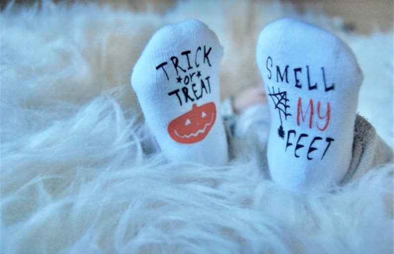 May include: A pair of white baby socks with Halloween-themed designs. One sock says "Trick or Treat" with a pumpkin and the other says "Smell My Feet" with a spider web.