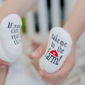 K&ouml;nnte beinhalten: Wei&szlig;e Babysocken mit schwarzem Text. Eine Socke sagt "Wenn du das lesen kannst..." und die andere Socke sagt "Bring mich zum Bauernhof!" mit einer roten Scheunengrafik.