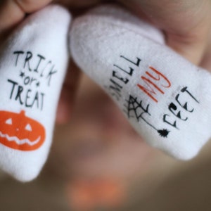 May include: A pair of white baby socks with Halloween-themed designs. One sock says "Trick or Treat" with a jack-o'-lantern and the other says "Smell My Feet" with a spider web.