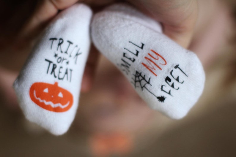 May include: A pair of white baby socks with Halloween-themed designs. One sock says "Trick or Treat" with a jack-o'-lantern and the other says "Smell My Feet" with a spider web.