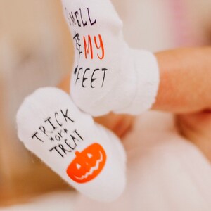May include: A pair of white baby socks with Halloween-themed designs. One sock says "Trick or Treat" with a pumpkin graphic, and the other says "Smell My Feet".