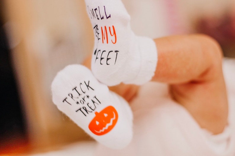 May include: A pair of white baby socks with Halloween-themed designs. One sock says "Trick or Treat" with a pumpkin graphic, and the other says "Smell My Feet".