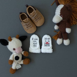K&ouml;nnte beinhalten: Ein Paar braune Babyschuhe, ein geh&auml;keltes braunes und wei&szlig;es Pferd, eine geh&auml;kelte wei&szlig;e und schwarze Kuh und zwei wei&szlig;e Baby-Sch&uuml;hchen mit dem Text "Wenn du das lesen kannst" und "Bring mich zum Bauernhof!" darauf.