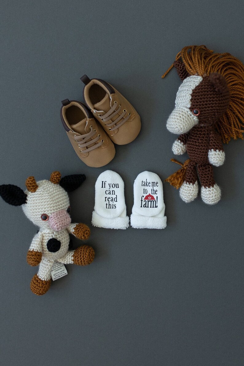 K&ouml;nnte beinhalten: Ein Paar braune Babyschuhe, ein geh&auml;keltes braunes und wei&szlig;es Pferd, eine geh&auml;kelte wei&szlig;e und schwarze Kuh und zwei wei&szlig;e Baby-Sch&uuml;hchen mit dem Text "Wenn du das lesen kannst" und "Bring mich zum Bauernhof!" darauf.