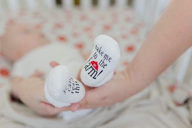 K&ouml;nnte beinhalten: Ein Paar wei&szlig;e Babyschuhe mit schwarzem Text. Ein Sch&uuml;hchen sagt "Nimm mich mit auf den Bauernhof!" mit einem roten Scheunen-Symbol. Das andere Sch&uuml;hchen sagt "Ich kann das lesen".